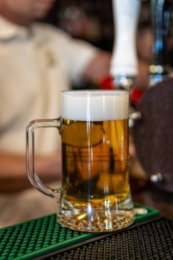 Kehribar birası ve beyaz köpükle dolu cam bardağa yakın plan çekilmiş. Arka planda odakları bozuk bir barmen var..