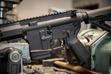 Black AR-15 style rifle receiver close-up on industrial metal machinery in a workshop setting.