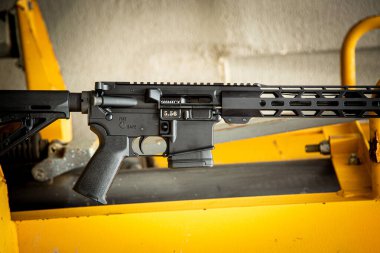 Black modern AR-15 style rifle assembly resting horizontally on bright yellow industrial machinery.