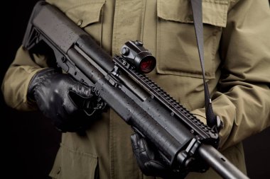 Man in tactical gloves and olive jacket holding a black pump-action shotgun with a red dot sight.