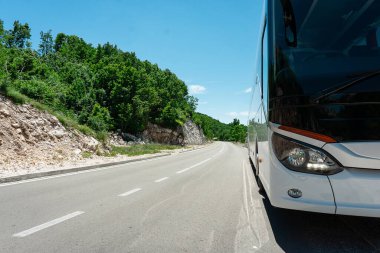 Beyaz bir tur otobüsü yeşil ağaçlarla kaplı güneşli, dolambaçlı bir dağ yolunda park halindedir..