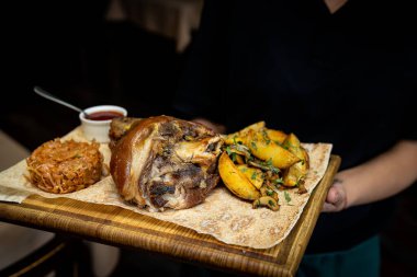 Garson, ahşap bir tahtanın üzerinde patates ve lahana ile kızarmış domuz budu servis ediyor..