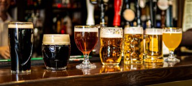 A variety of beer glasses and mugs with different colors of beer lined up on a wooden bar.