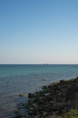 Kaya kıyısı, bulutsuz açık gökyüzü, ufukta iki römorkör.