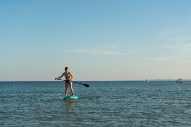 12 yaşında bir çocuk kıyıya yakın bir denizde SUP tahtasının üzerinde durmayı öğreniyor..