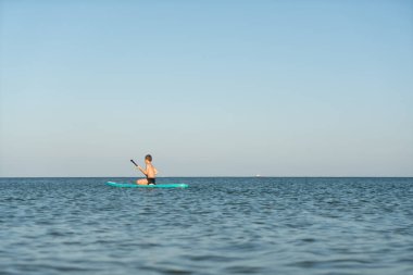 12 yaşında bir çocuk kıyıya yakın bir denizde SUP tahtasının üzerinde durmayı öğreniyor..