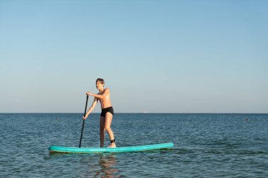12 yaşında bir çocuk kıyıya yakın bir denizde SUP tahtasının üzerinde durmayı öğreniyor..