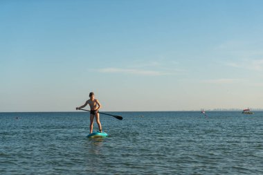 12 yaşında bir çocuk kıyıya yakın bir denizde SUP tahtasının üzerinde durmayı öğreniyor..