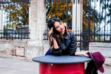 Kırmızı ve sarı sonbahar arkaplanındaki modacı kadın, Tasasız genç kadını modaya uygun bir şekilde parka bırakır ve güler durur. Kız güz gününde poz veriyor, güzel havanın tadını çıkarıyor..