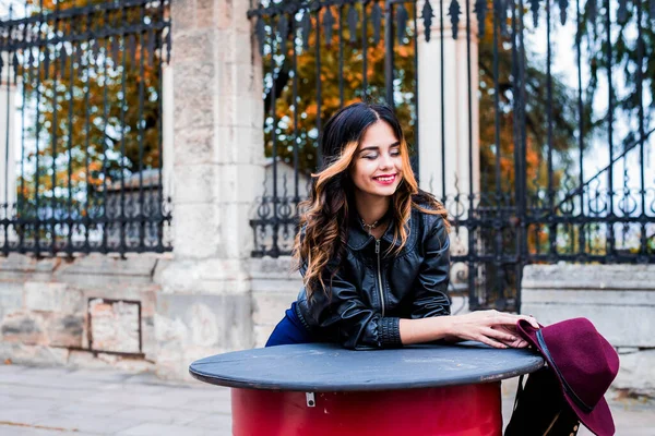 Uzun saçlı, kırmızı dudaklı, gülen güzel bir kız. Sonbahar caddesinde şık bir şapka takıyor. Açık hava portresi, gün ışığı. Kadın sonbahar moda konsepti. Kırmızı ve sarı sonbahar yapraklarının arka planında moda kadını