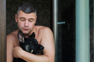 Pencerenin arka planında kameraya bakan mutlu yaşlı adamın yakın plan portresi. Doğu Avrupa çoban köpeğini kucaklayan adam. Gülümsüyor, iyi günlerin tadını çıkarıyor ve evcil hayvanlarla poz veriyor..