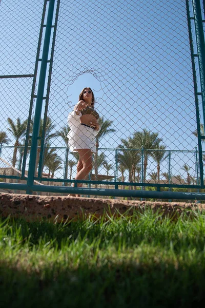 Stylish cute teenage model dressed in summer hipster vintage white stylish clothes. Trendy girl posing in sunglasses girl in hole behind fence of the lattice at the sports field on a summer sunny day.