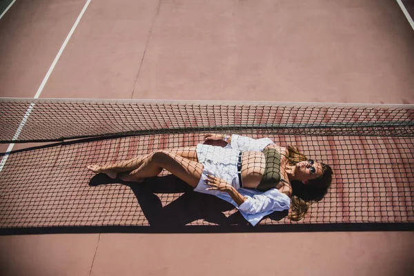 时髦可爱的少女模特穿着夏令时髦的老式白色时尚服装 一个穿着太阳镜的时髦姑娘躺在网球场上 最初的阴影来自运动场的网格 — 图库照片