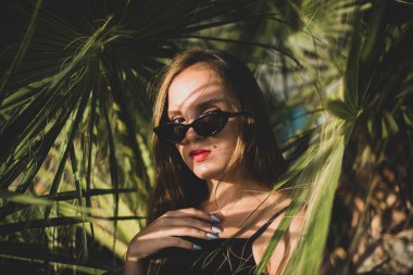 Güneş gözlüğü takan kadın palmiye ağacıyla poz veriyor. Yüzünde palmiye yaprağı gölgeleri olan güzel bir genç kızın portresi.