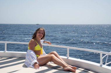 Neşeli genç kadın portresi. Yelkenli yatın güvertesinde oturan mutlu genç kız, yaz gezisinde tropikal denizde adaları keşfederken eğleniyor. Seyahat macerası, yat gezisi.