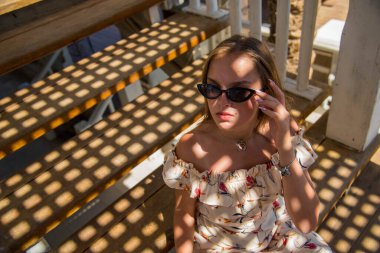 Güneşli bir yaz gününde plajdaki kafede güneş gözlüğü takan güzel bir kızın moda portresi. Genç, zayıf, güzel kadın yaz tatili, güneşli..