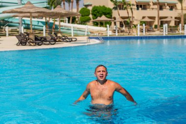 Yakışıklı, yarı çıplak, neşeli ve gülümseyen bir adam mavi su havuzunda gülüyor. Yaz manzarasında yüzme havuzunda gülümseyen yakışıklı bir adamın fotoğrafı.