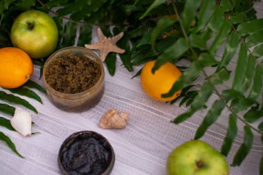 Ev yapımı kozmetik malzemelerinin resmi. Aroma temalı. Siyah maske, kil. Bitki özleriyle organik kozmetik ürünler beyaz yapraklı yeşil arka planda elma, limon ve portakallar