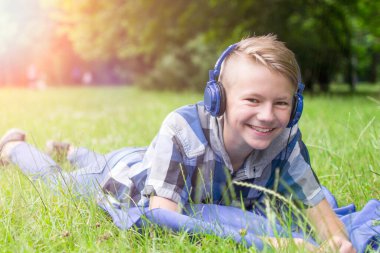 Dışarıda kulaklıkları olan 12-16 yaşlarında gülen bir çocuk. Çocukluk. Öğrenci çocuk. Çimlerin üzerinde uzanarak, yaz.