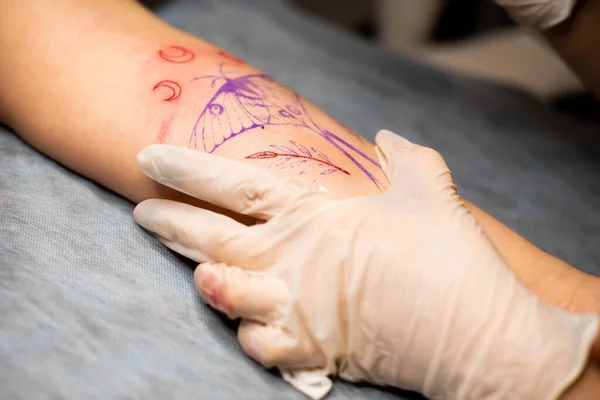 Master tattoo draws the red paint on the clients tattoo. Tattoo artist holding a pink tattoo machine in black sterile gloves and working on the professional blue mat.