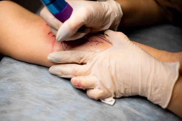 Master tattoo draws the red paint on the clients tattoo. Tattoo artist holding a pink tattoo machine in black sterile gloves and working on the professional blue mat.