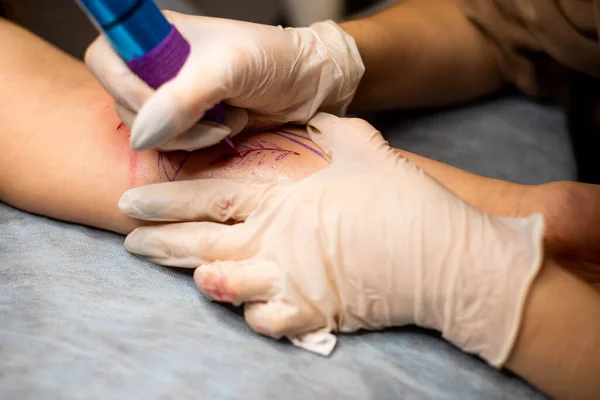 Master tattoo draws the red paint on the clients tattoo. Tattoo artist holding a pink tattoo machine in black sterile gloves and working on the professional blue mat.