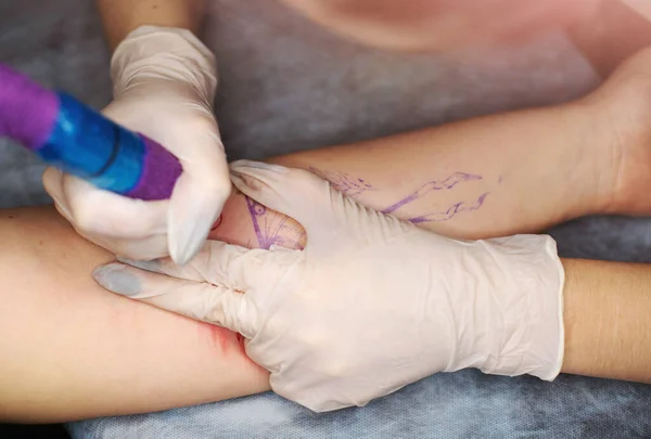 Master tattoo draws the red paint on the clients tattoo. Tattoo artist holding a pink tattoo machine in black sterile gloves and working on the professional blue mat.