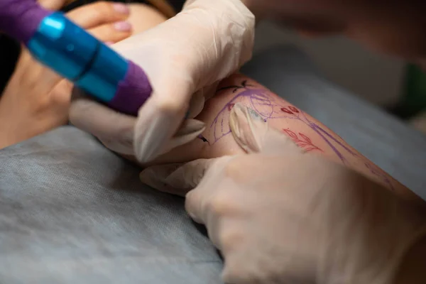 Master tattoo draws the red paint on the clients tattoo. Tattoo artist holding a pink tattoo machine in black sterile gloves and working on the professional blue mat.