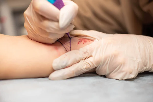 Master tattoo draws the red paint on the clients tattoo. Tattoo artist holding a pink tattoo machine in black sterile gloves and working on the professional blue mat.