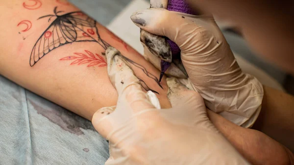 Master tattoo draws the red paint on the clients tattoo. Tattoo artist holding a pink tattoo machine in black sterile gloves and working on the professional blue mat.