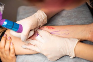 Ana dövme müşterilerin dövmelerindeki kırmızı boyayı çizer. Siyah steril eldivenli pembe dövme makinesi tutan dövme sanatçısı profesyonel mavi paspas üzerinde çalışıyor..