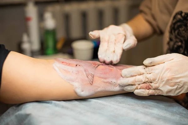 Master tattoo draws the red paint on the clients tattoo. Tattoo artist holding a pink tattoo machine in black sterile gloves and working on the professional blue mat.