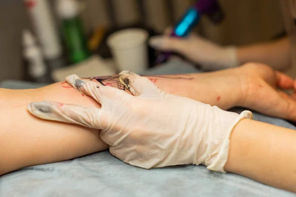 Master tattoo draws the red paint on the clients tattoo. Tattoo artist holding a pink tattoo machine in black sterile gloves and working on the professional blue mat.