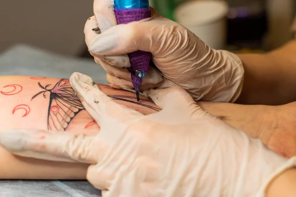 Master tattoo draws the red paint on the clients tattoo. Tattoo artist holding a pink tattoo machine in black sterile gloves and working on the professional blue mat.