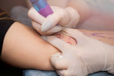 Ana dövme müşterilerin dövmelerindeki kırmızı boyayı çizer. Siyah steril eldivenli pembe dövme makinesi tutan dövme sanatçısı profesyonel mavi paspas üzerinde çalışıyor..