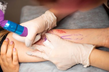 Ana dövme müşterilerin dövmelerindeki kırmızı boyayı çizer. Siyah steril eldivenli pembe dövme makinesi tutan dövme sanatçısı profesyonel mavi paspas üzerinde çalışıyor..