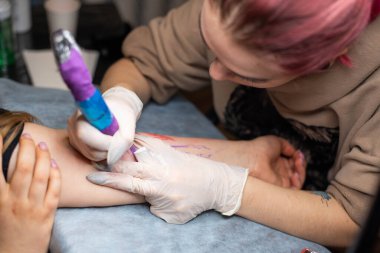 Ana dövme müşterilerin dövmelerindeki kırmızı boyayı çizer. Siyah steril eldivenli pembe dövme makinesi tutan dövme sanatçısı profesyonel mavi paspas üzerinde çalışıyor..