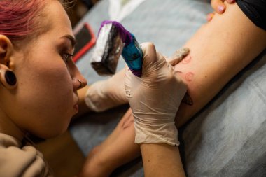 Ana dövme müşterilerin dövmelerindeki kırmızı boyayı çizer. Siyah steril eldivenli pembe dövme makinesi tutan dövme sanatçısı profesyonel mavi paspas üzerinde çalışıyor..