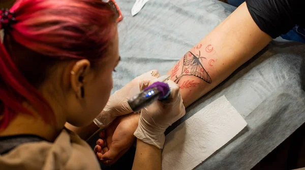 Master tattoo draws the red paint on the clients tattoo. Tattoo artist holding a pink tattoo machine in black sterile gloves and working on the professional blue mat.