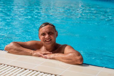 Yakışıklı, yarı çıplak, neşeli ve gülümseyen bir adam mavi su havuzunda gülüyor. Yaz manzarasında yüzme havuzunda gülümseyen yakışıklı bir adamın fotoğrafı.