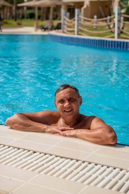 Yakışıklı, yarı çıplak, neşeli ve gülümseyen bir adam mavi su havuzunda gülüyor. Yaz manzarasında yüzme havuzunda gülümseyen yakışıklı bir adamın fotoğrafı.