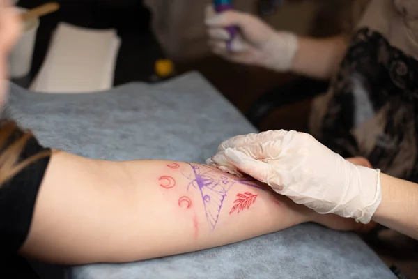 Master tattoo draws the red paint on the clients tattoo. Tattoo artist holding a pink tattoo machine in black sterile gloves and working on the professional blue mat.