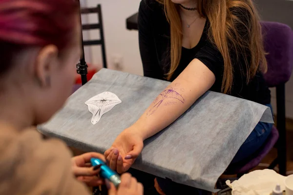 Charming girl with pink hair and tattoos stuffs a tattoo on a woman\'s hand in a tattoo parlor, creative small business and tattoo master. Hipster woman in gloves draws a tattoo