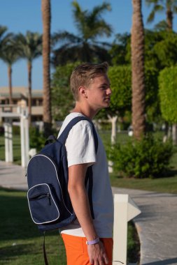 Yakışıklı genç çocuk sahilde kırmızı tişörtlü palmiye ağaçlarının altında poz veriyor ve eğleniyor. Moda çocuğu poz veriyor. Güneş gözlüklü komik ve pozitif adam.