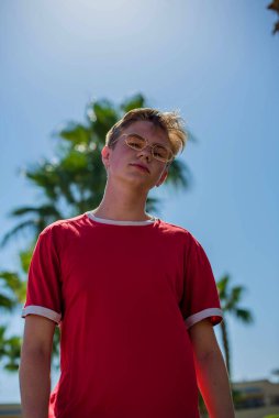Yakışıklı genç çocuk sahilde kırmızı tişörtlü palmiye ağaçlarının altında poz veriyor ve eğleniyor. Moda çocuğu poz veriyor. Güneş gözlüklü komik ve pozitif adam.