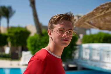 Yakışıklı genç çocuk sahilde kırmızı tişörtlü palmiye ağaçlarının altında poz veriyor ve eğleniyor. Moda çocuğu poz veriyor. Güneş gözlüklü komik ve pozitif adam.