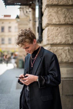 Sokak tarzı kıyafetler giymiş, boynuna zincirler takmış yürüyüş için telefon takmış pozitif erkek kıvırcık saçlı. Gençlik ve yaşam tarzı kavramı.
