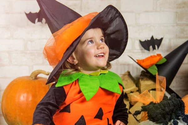 The concept of Halloween, funny face. Funny child girl in pumpkin costume for Halloween with pumpkins and holiday attributes. Children\'s party. Cute little witch dressed in hat with a pumpkin.