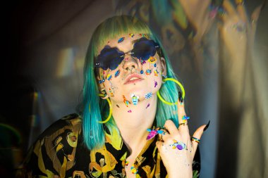 Yakın plan moda portresi yeşil ve mavi saçlı güzel bir kız. Yüzüne komik emoji çıkartmaları yapıştırılmış lüks profesyonel makyajlı güzel bir moda kızı. Genç kadın güneş gözlüğü