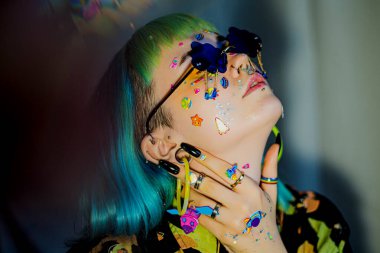 Yakın plan moda portresi yeşil ve mavi saçlı güzel bir kız. Yüzüne komik emoji çıkartmaları yapıştırılmış lüks profesyonel makyajlı güzel bir moda kızı. Genç kadın güneş gözlüğü